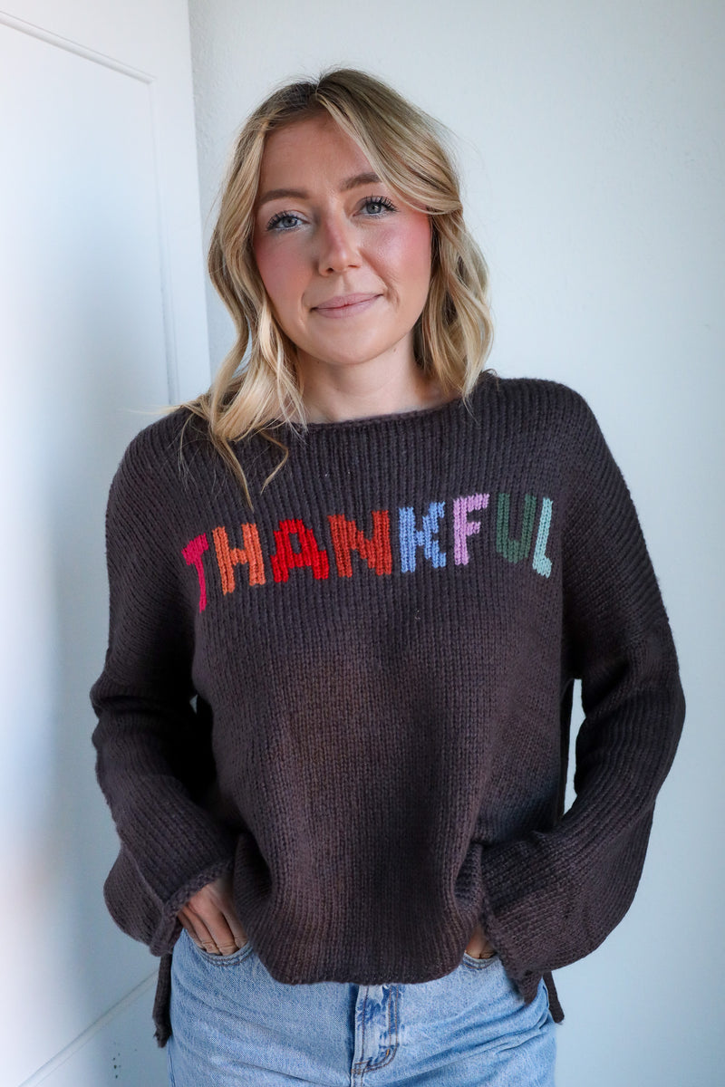 Woman wearing a sweater with 'THANKFUL' in colorful letters against a white wall.