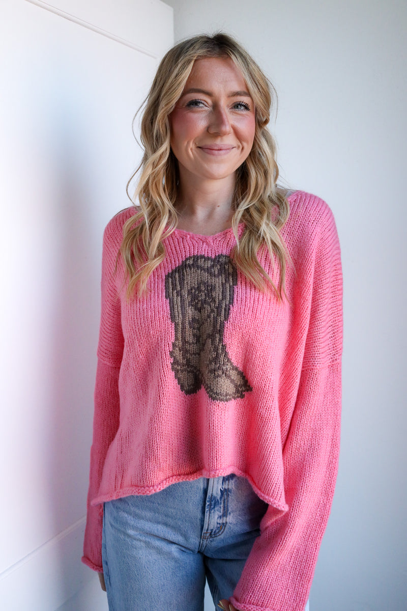 Woman wearing a pink sweater with a graphic design, standing against a white wall.