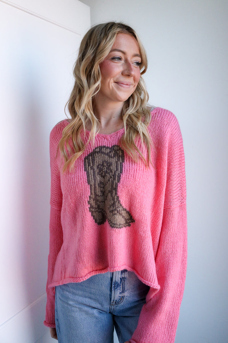 Woman wearing a pink sweater with a cowgirl boot design against a white wall.