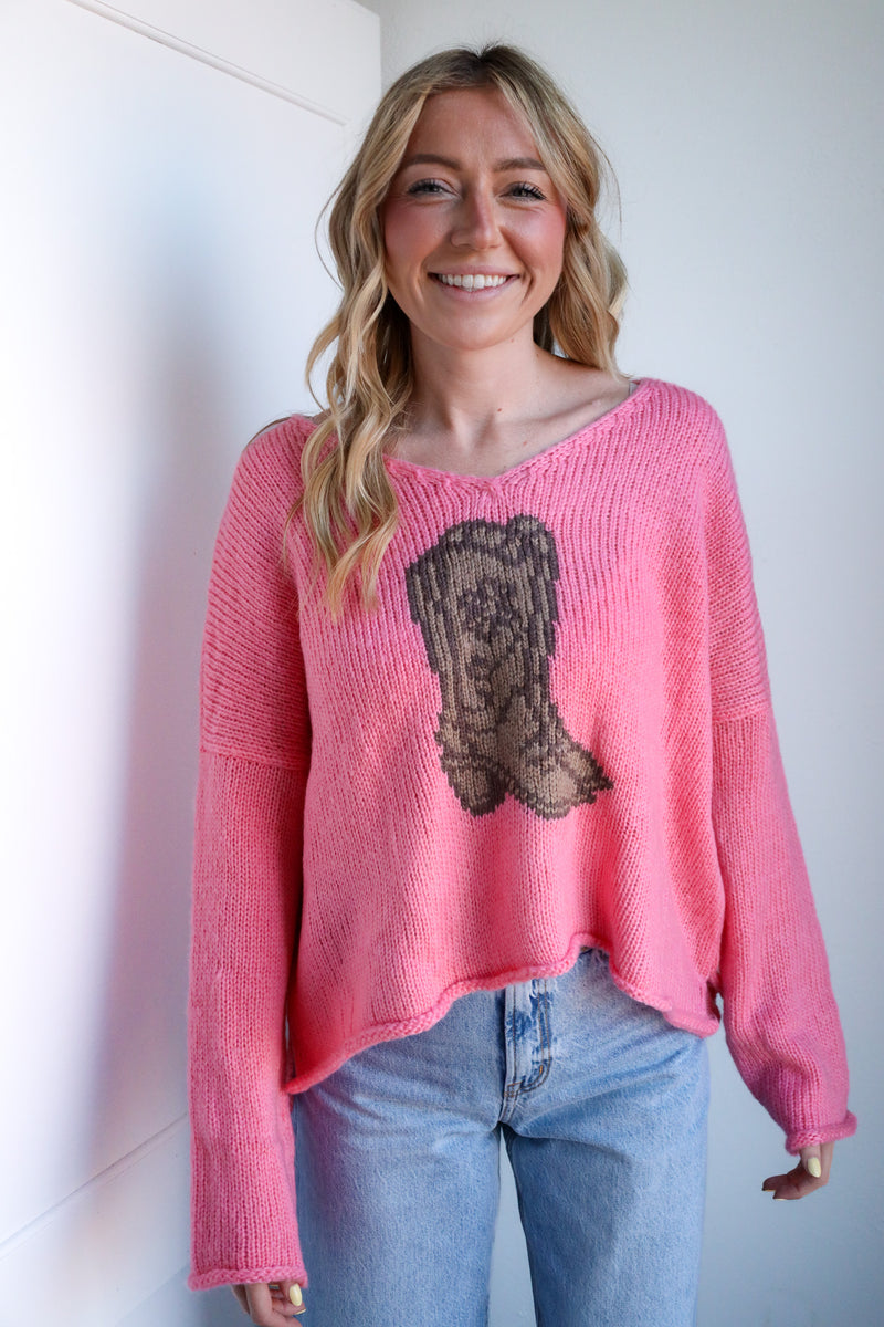 Woman wearing a pink sweater with a cowboy boot design against a white wall.