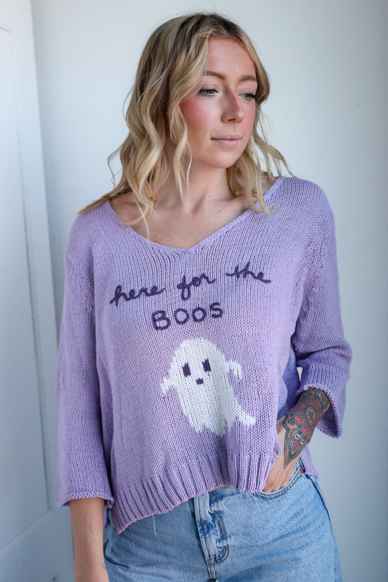 Woman wearing a purple sweater with a ghost design and text, standing against a plain background.