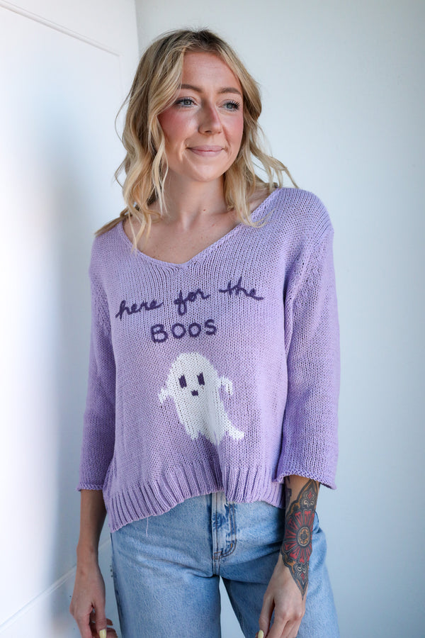 Woman wearing a purple sweater with a ghost design and text, standing against a plain background.
