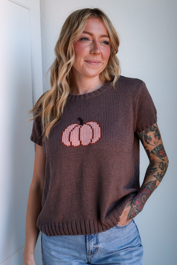 Woman wearing a brown sweater with pink pumpkin design against a white wall.