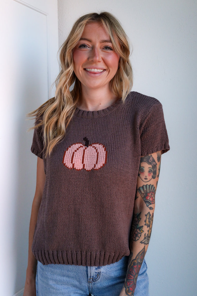 Woman wearing a brown sweater with pink pumpkin design against a white wall.