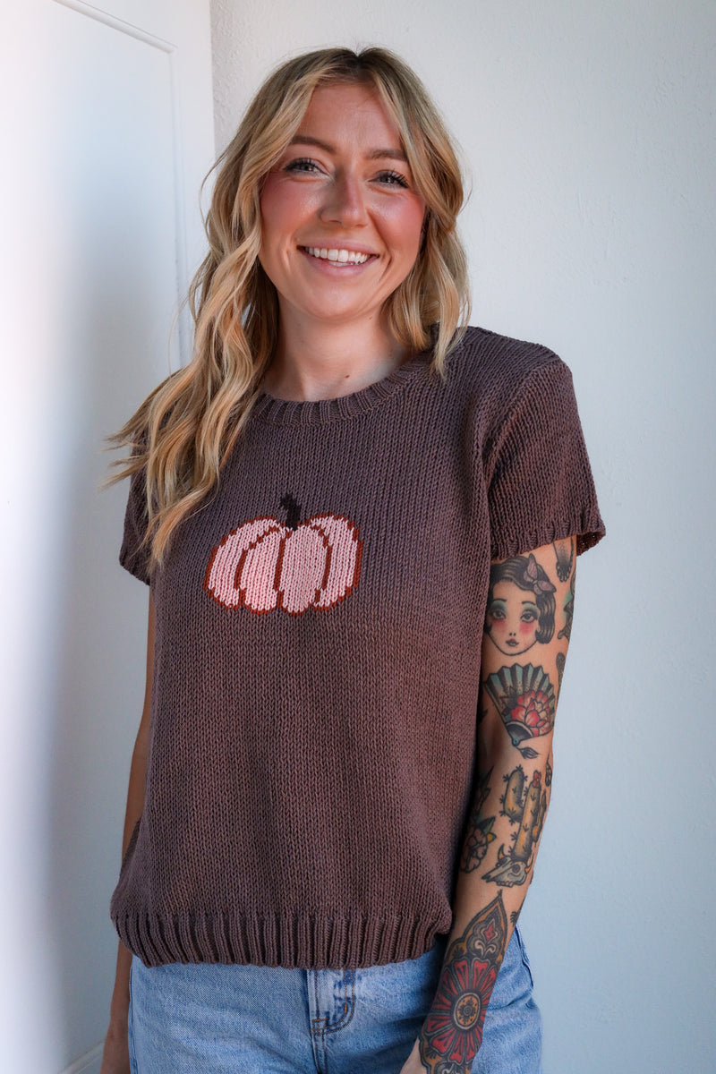 Woman wearing a brown sweater with a pink pumpkin design against a white wall.