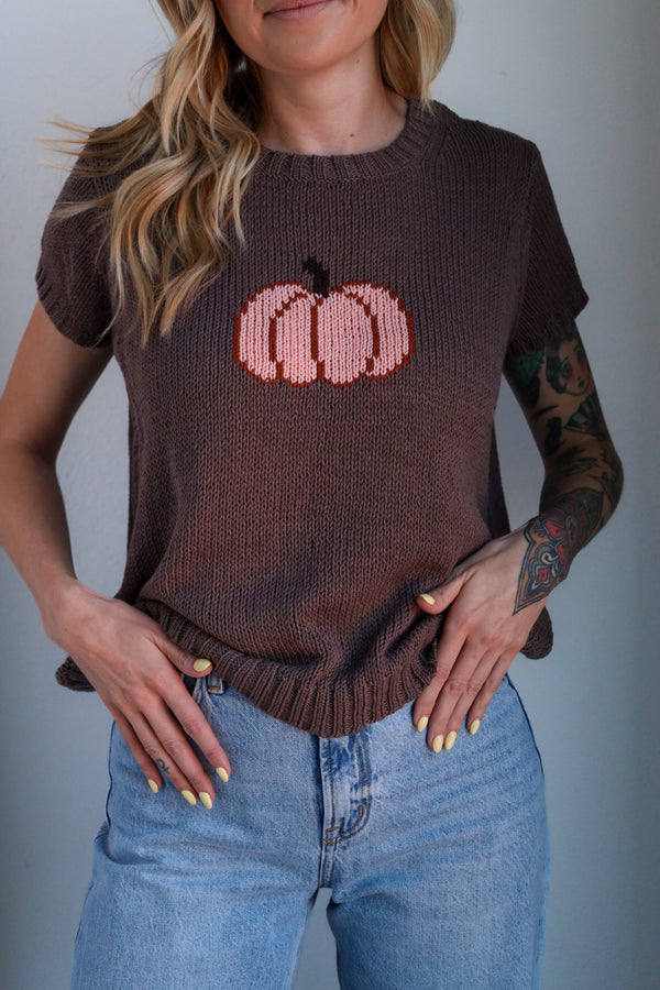 Person wearing a brown sweater with a pink pumpkin design and blue jeans against a gray background