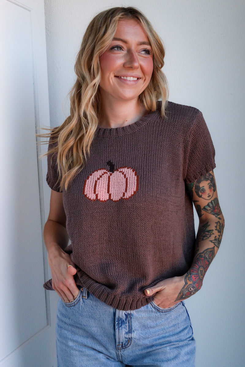 Woman wearing a brown sweater with pink pumpkin design against a white wall