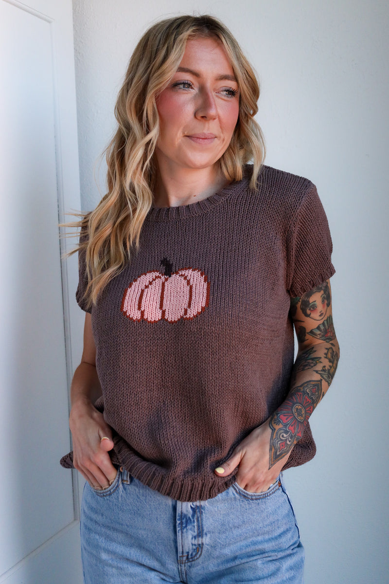 Woman wearing a brown sweater with pink pumpkin design against a white wall
