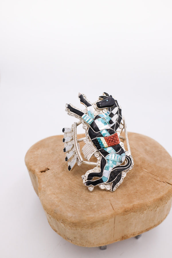 Today we have this stunning sterling silver horse Zuni Pony cuff bracelet with an Acoma black jet Zuni Pony centerpiece measuring 4 inches long. This incredible Acoma black jet horse is adorned and inlaid with turquoise, red spiny oyster, white shell, yellow shell and abalone. Bracelet is placed on a wooden display.