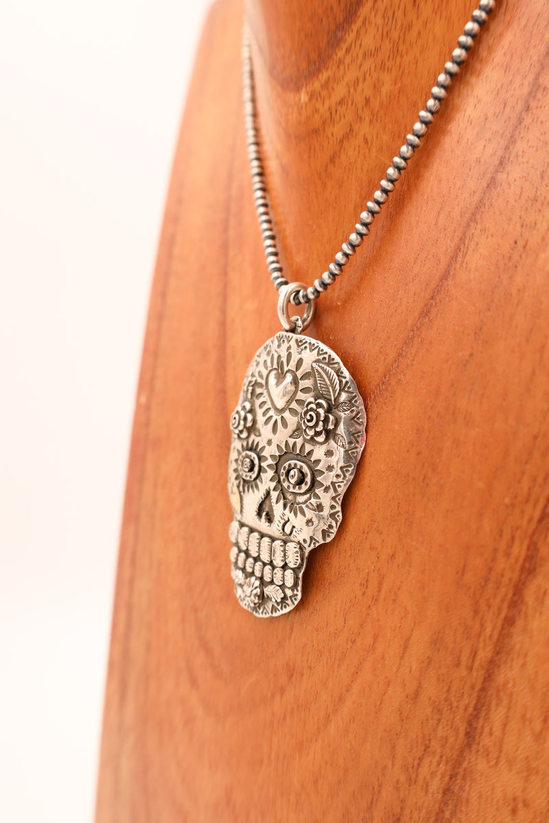 Sterling silver sugar skull with hand stamped details and a 14K gold heart in the center of the head handing on a display necklace hanging on a wooden neck display