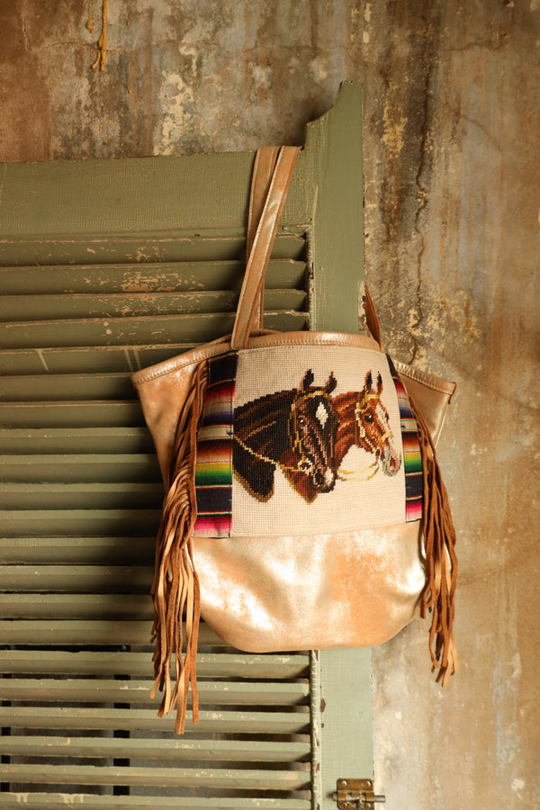 Introducing the Totem 2 Horses Serape Leather Tote - a one-of-a-kind bag that exudes style and uniqueness. Made from metallic cream leather, this tote features fringe and exquisite embroidery of two horse heads with a vibrant serape pattern on either side. Make a statement with this stunning tote!