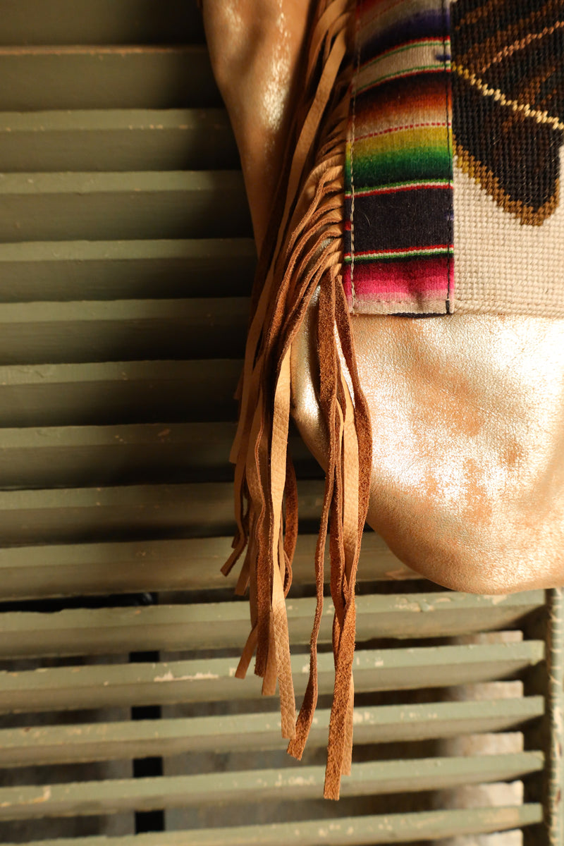 Introducing the Totem 2 Horses Serape Leather Tote - a one-of-a-kind bag that exudes style and uniqueness. Made from metallic cream leather, this tote features fringe and exquisite embroidery of two horse heads with a vibrant serape pattern on either side. Make a statement with this stunning tote!