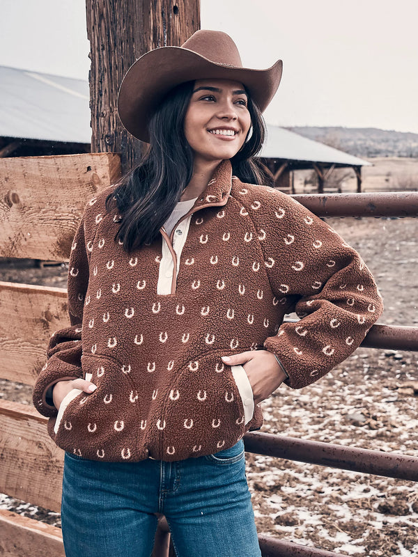 Woman wearing a brown patterned sweater and cowboy hat standing in a rustic outdoor setting.