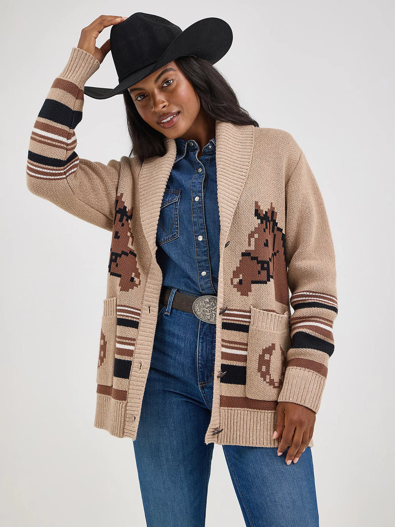 Woman wearing a beige cardigan with horse patterns, blue jeans, and a black cowboy hat on a white background.