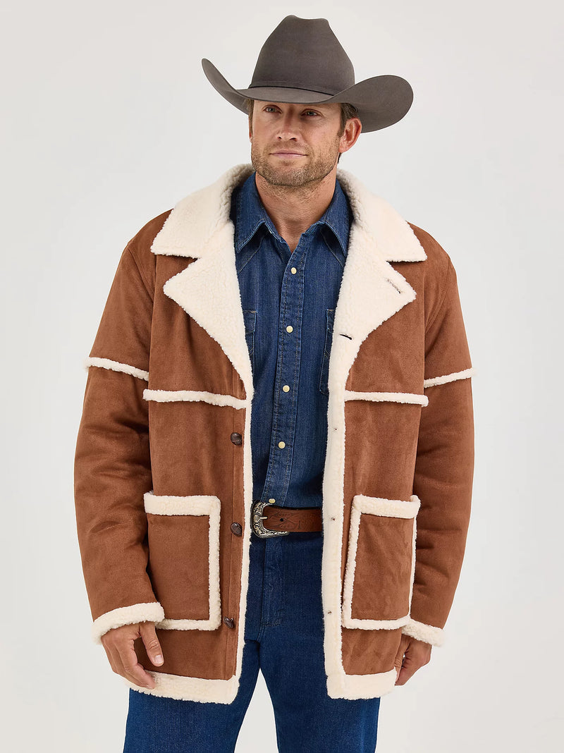 Man wearing a brown shearling jacket with white fur trim, blue jeans, and a cowboy hat on a white background.