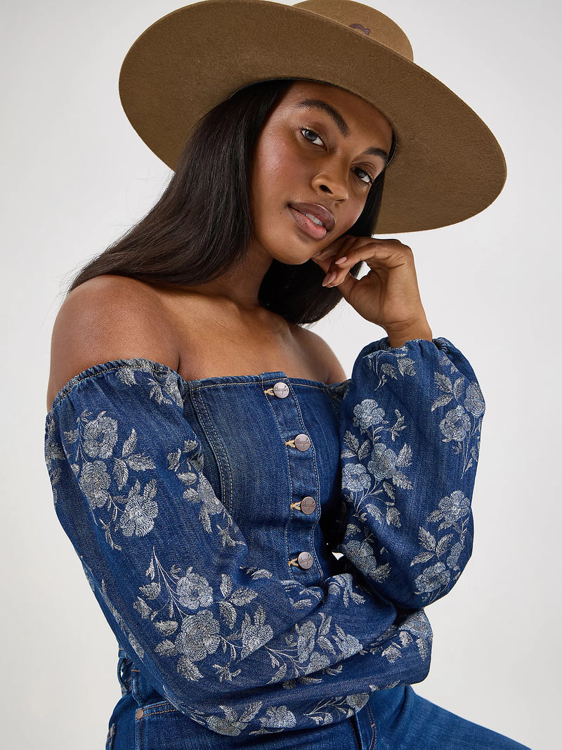 Woman wearing a blue floral off-shoulder top and brown hat against a white background