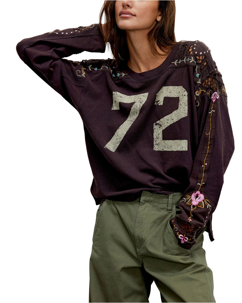 Person wearing a dark purple sweatshirt with the number 72 and floral patterns, paired with green pants on a white background.