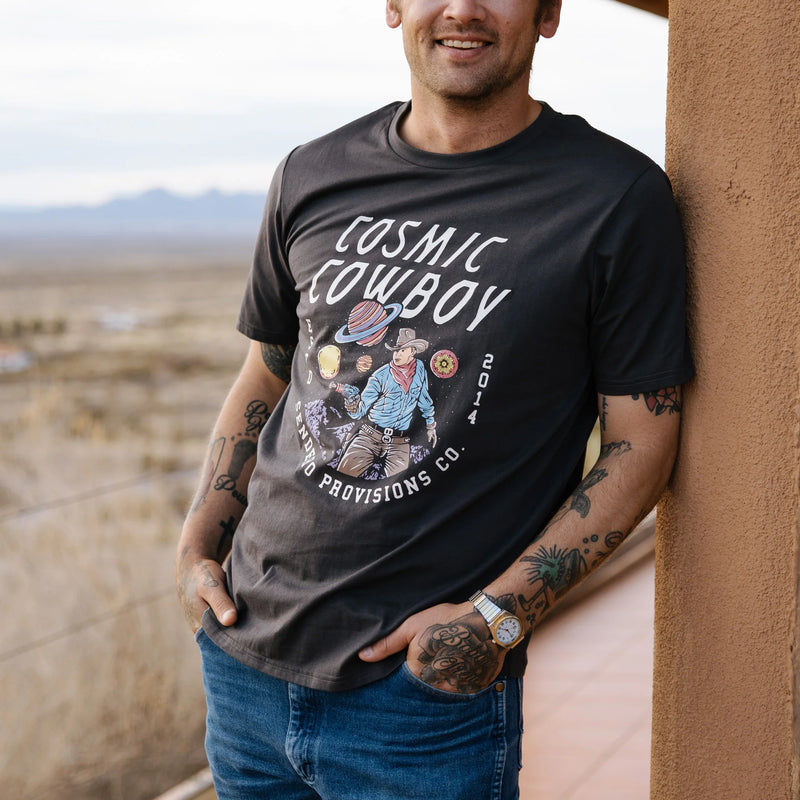 Man wearing black short sleeve shirt with script "cosmic cowboy ESTD 2014 Sendero Provisions Co." on front.