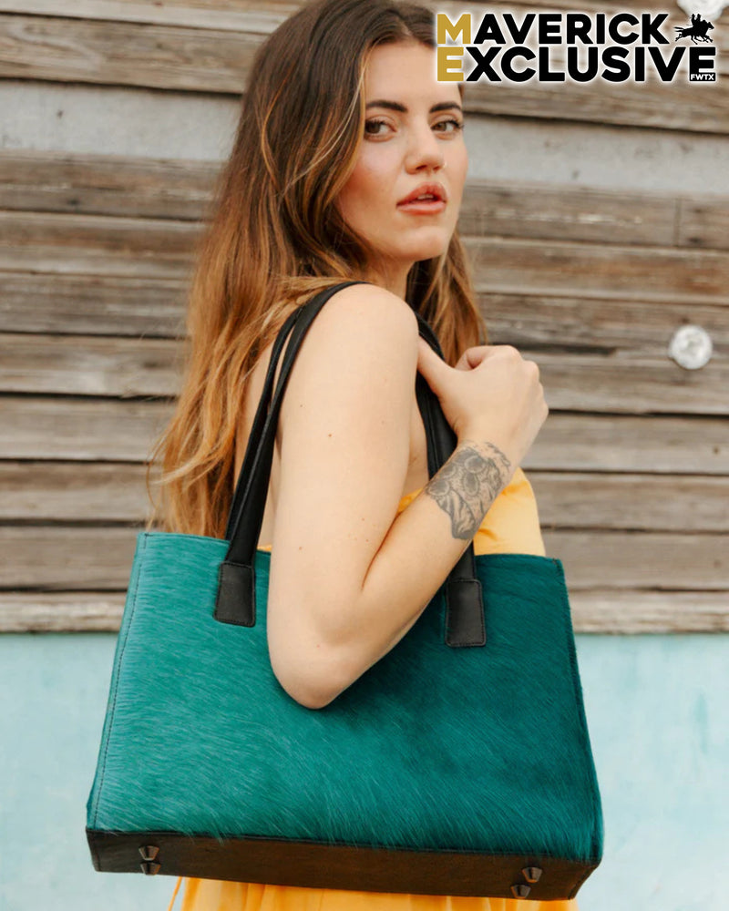 Woman holding a teal tote bag against a wooden wall, with 'Maverick Exclusive' branding.