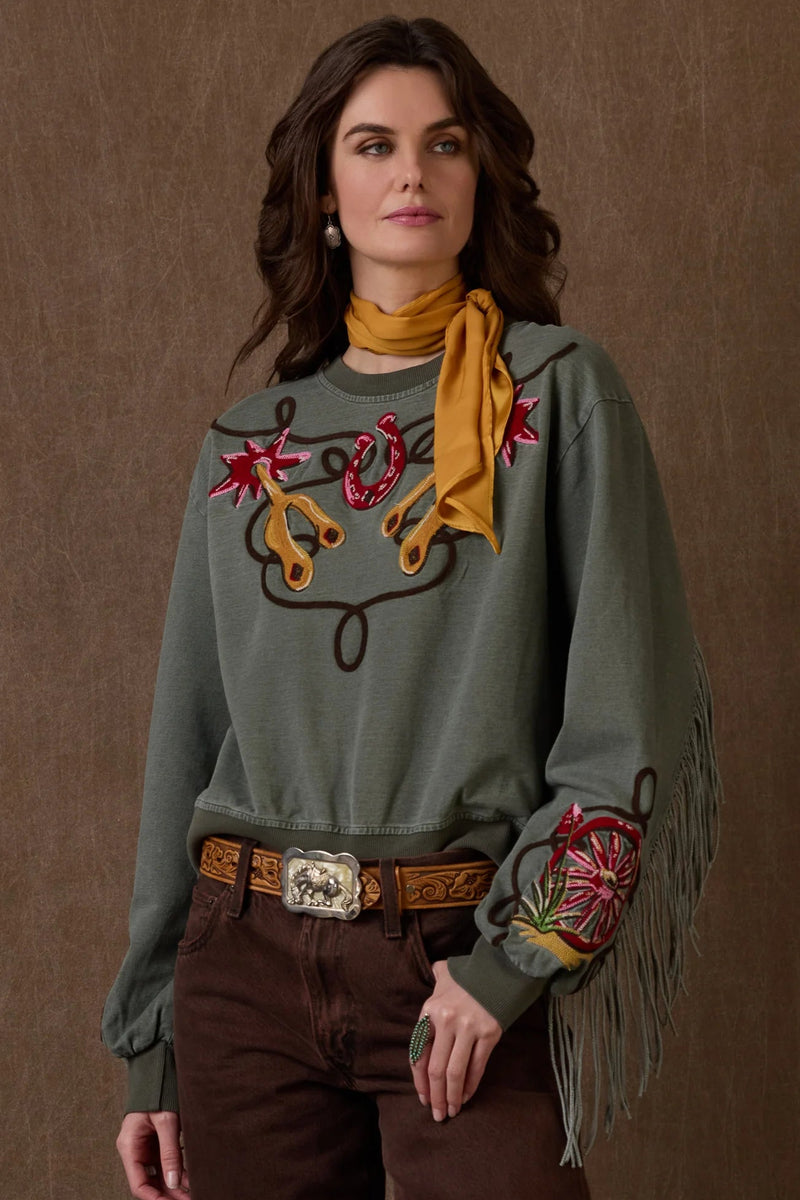 Woman wearing a green embroidered sweater with fringes and a yellow scarf against a brown background.