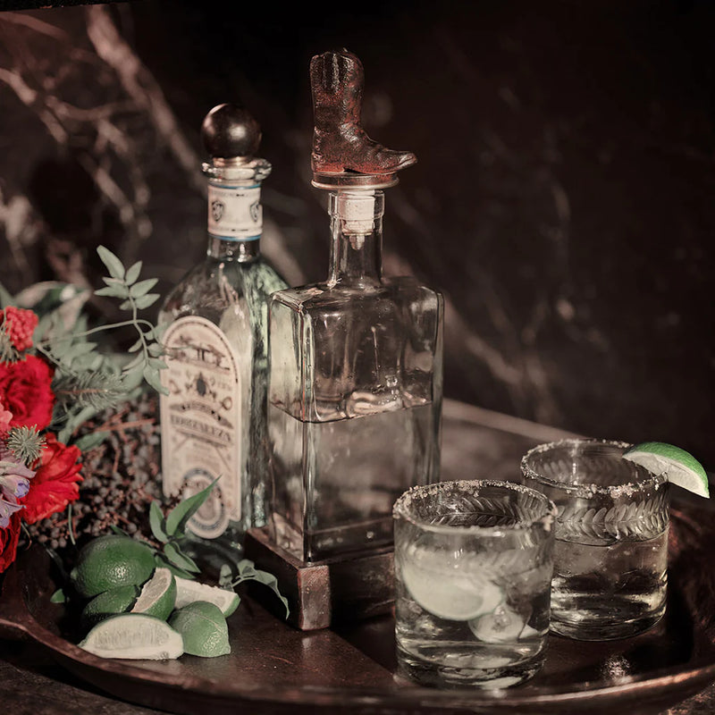 Two glass bottles with decorative elements on a wooden tray with glasses and limes.