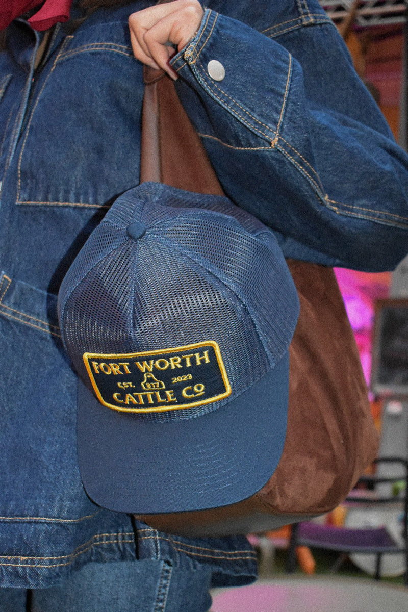 Blue cap with 'Fort Worth Cattle Co.' logo held by a person wearing a denim jacket.