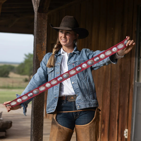 Woman holding a long, decorative belt in front of a wooden building