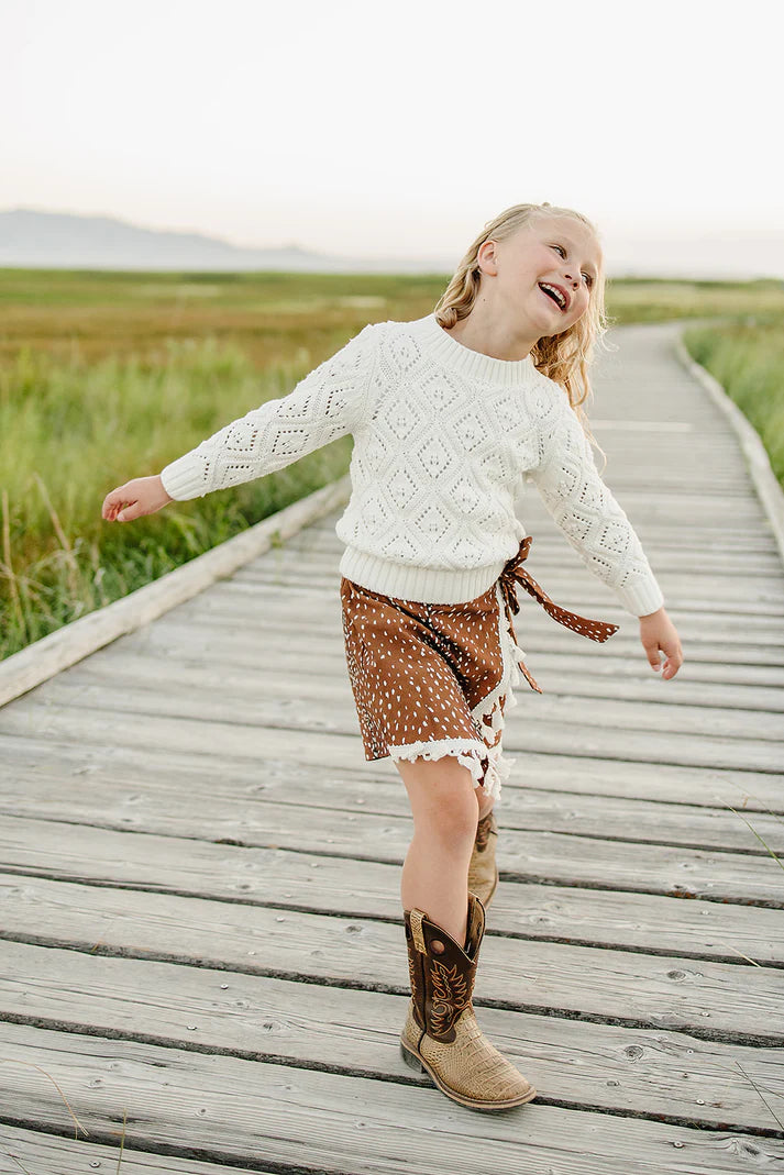 Child in a white sweater and brown patterned shorts walking on a wooden boardwalk with grassy fields in the background.