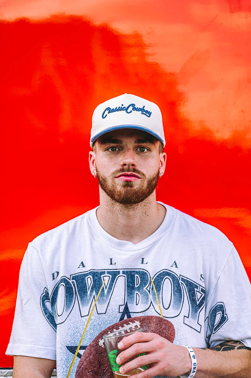 Man wearing a Dallas Cowboys cap and shirt, holding a football and drink against a red background
