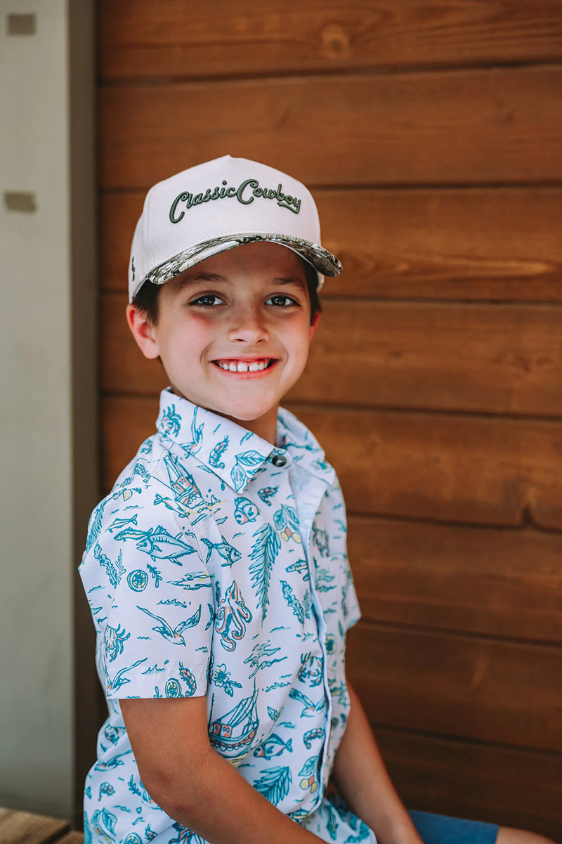 Young boy wearing a cap and patterned shirt against a wooden wall