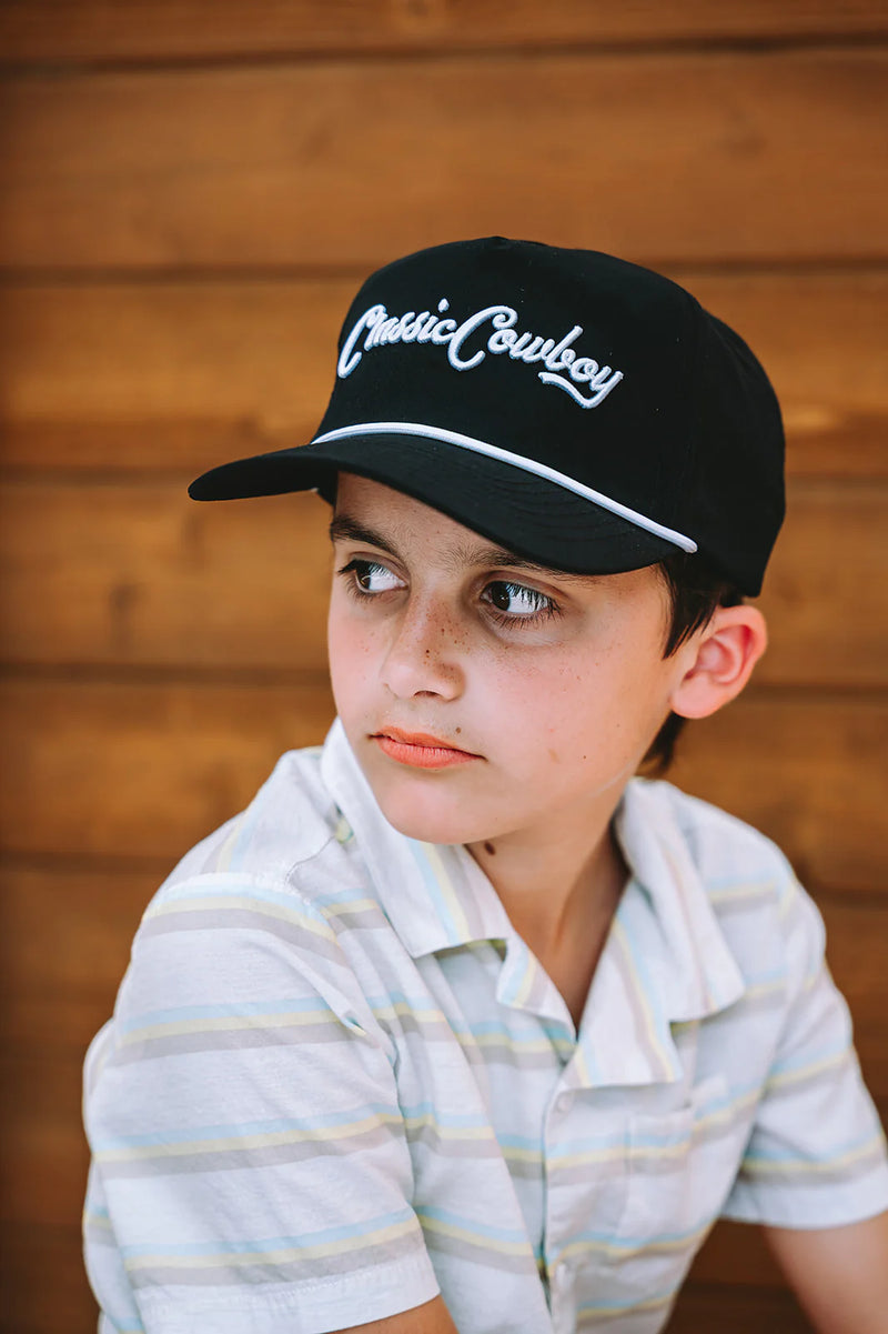 Child wearing a black cap with 'Classic Cowboy' text against a wooden background