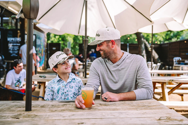 Two men sitting at an outdoor table with a beer, surrounded by umbrellas and other patrons.