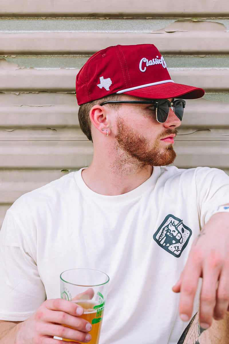 Man wearing a red cap with a logo, sunglasses, and holding a beer glass against a metallic background.