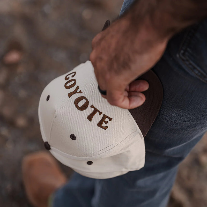Person holding a white cap with 'COYOTE' branding against a blurred natural background