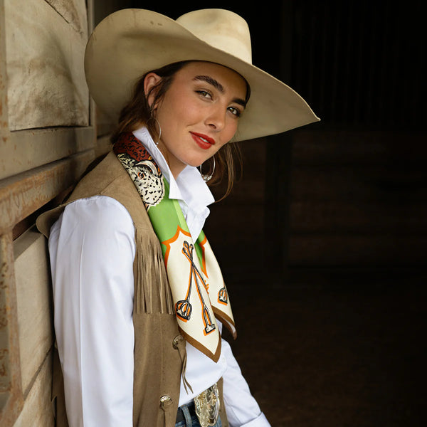 Woman wearing a cowboy hat and vest with a decorative scarf, leaning against a stone wall.