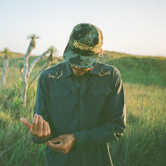 MAN WEARING DARK GREY SNAP BUTTON SHIRT WITH A SNAKE AND A MOON ON EITHER SHOULDER STANING IN A FIELD 