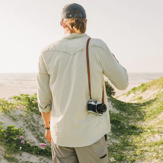 MAN WEARING GREEN BUTTON UP SHIRT WITH THIN STRIPES LOOKING AT THE OCEAN HOLDING A CAMERA