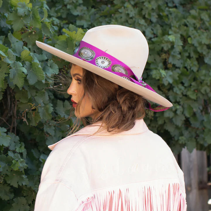 Woman wearing a white hat with a purple band and pink fringe jacket against a green leafy background
