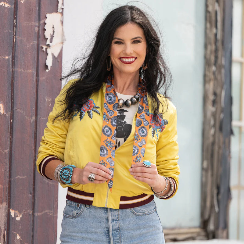 Woman wearing a yellow jacket with floral embroidery, denim jeans, and a colorful scarf.