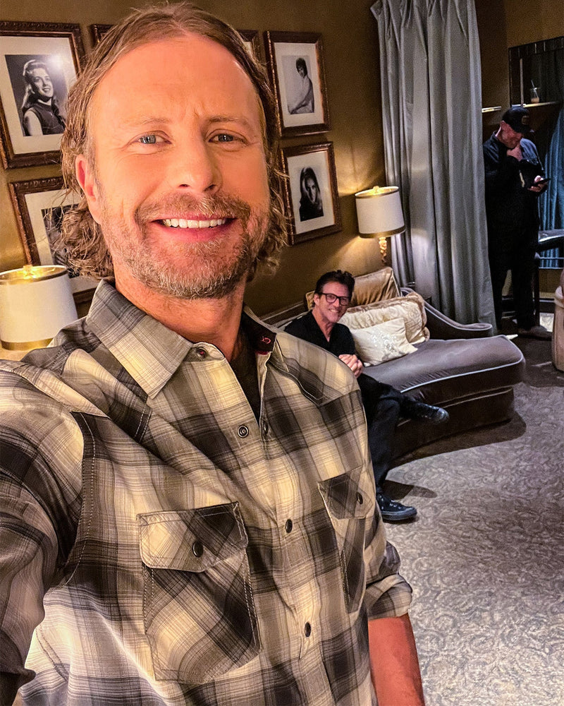 Man taking a selfie in a living room with another person sitting on a couch.