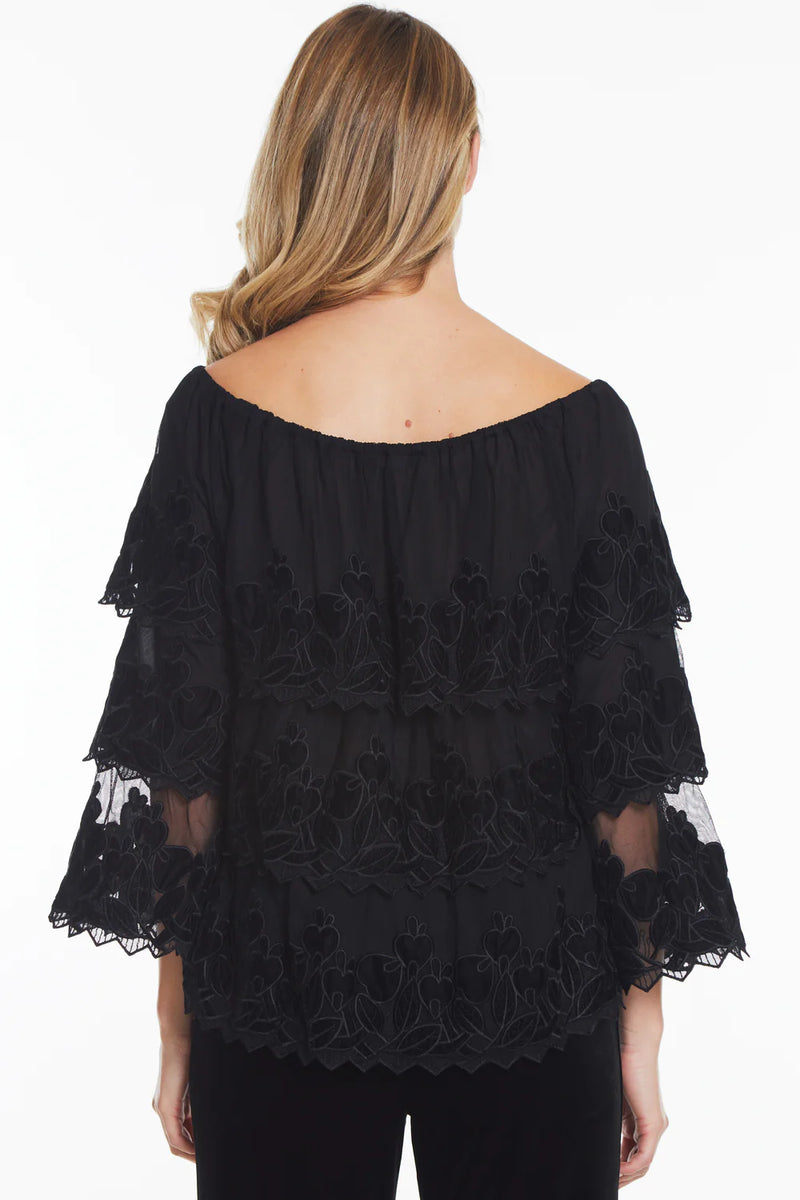 Woman wearing black tiered tunic top with long sleeves, black floral embellishment and off the shoulder detailing.