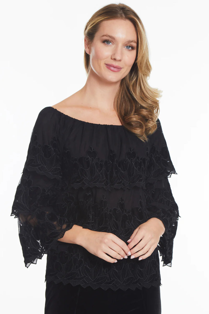 Woman wearing black tiered tunic top with long sleeves, black floral embellishment and off the shoulder detailing.
