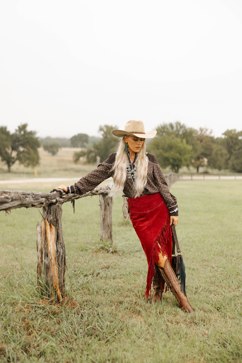 Woman in a red skirt and cowboy hat leaning against a wooden fence in a field.