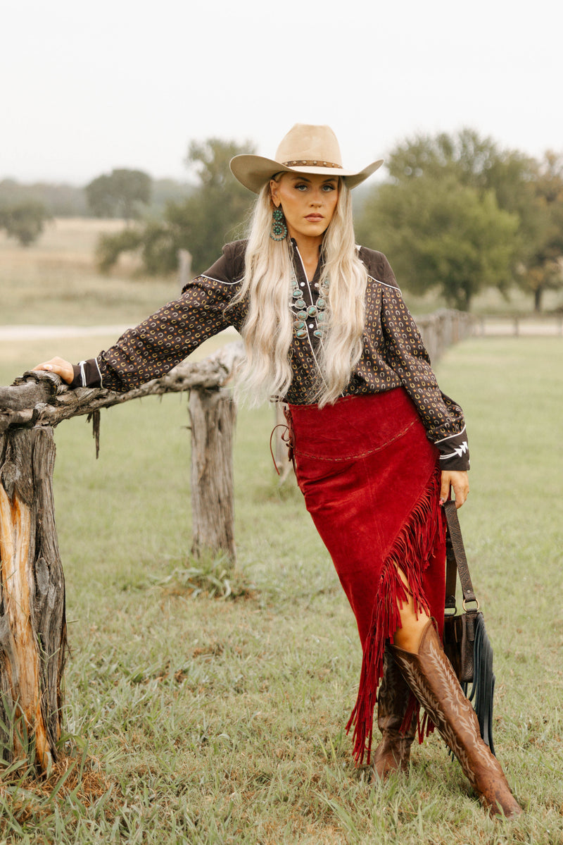 Woman in cowboy hat and red skirt standing by a wooden fence in a field