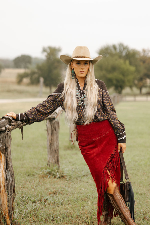 Woman in cowboy hat and red skirt standing by a wooden fence in a field.