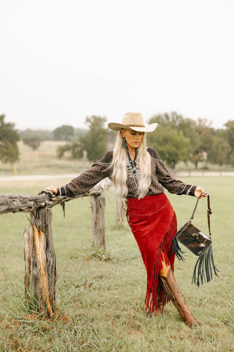Woman in cowboy hat and red skirt with fringe leaning against a wooden fence in a field.