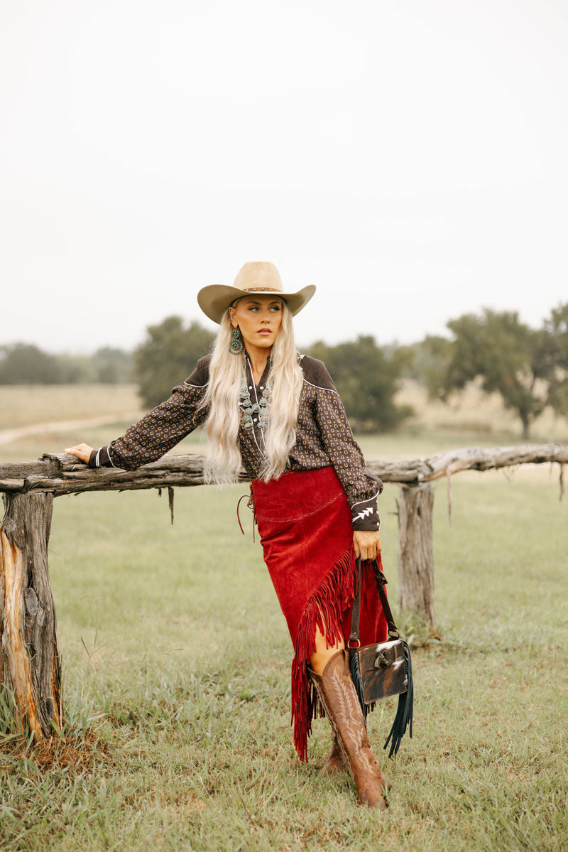 Woman in cowboy hat and red fringed skirt standing by a wooden fence in a field.