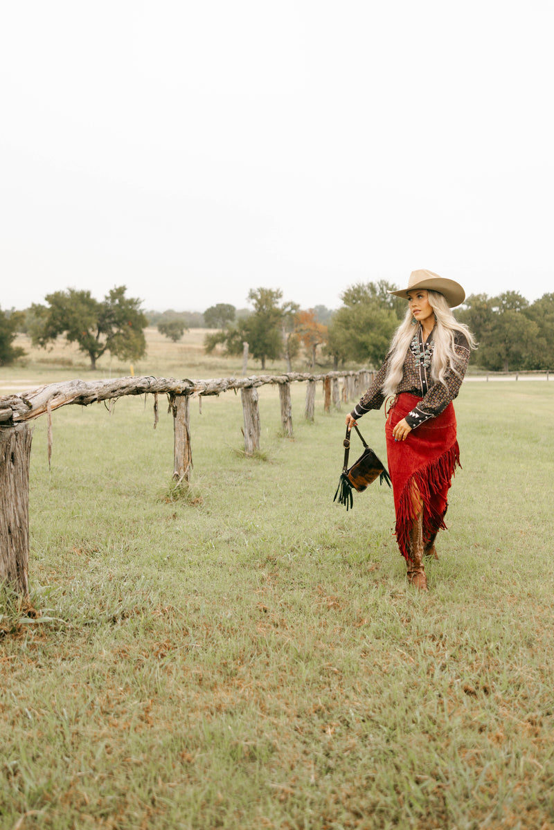 Woman in a red skirt and cowboy hat walking in a field with a wooden fence and trees in the background.