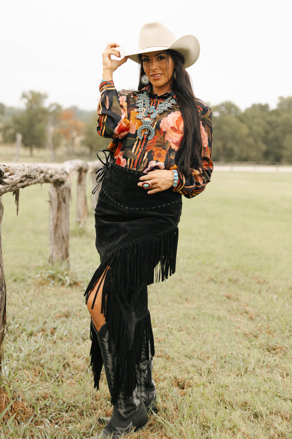Woman in a colorful patterned shirt and black fringed skirt standing in a field with a wooden fence.
