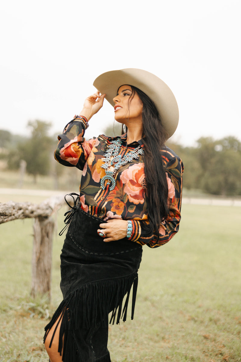 Woman in a floral blouse and black fringe skirt with a cowboy hat outdoors.
